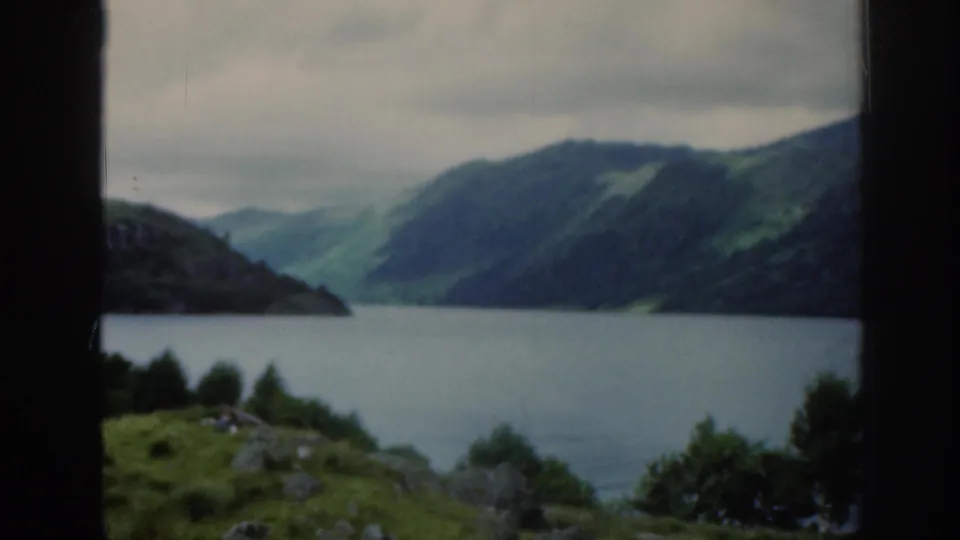 A scenic Scottish loch fills the frame, with dark mountain slopes, green patches, low trees, and soft 8mm color under grey clouds.