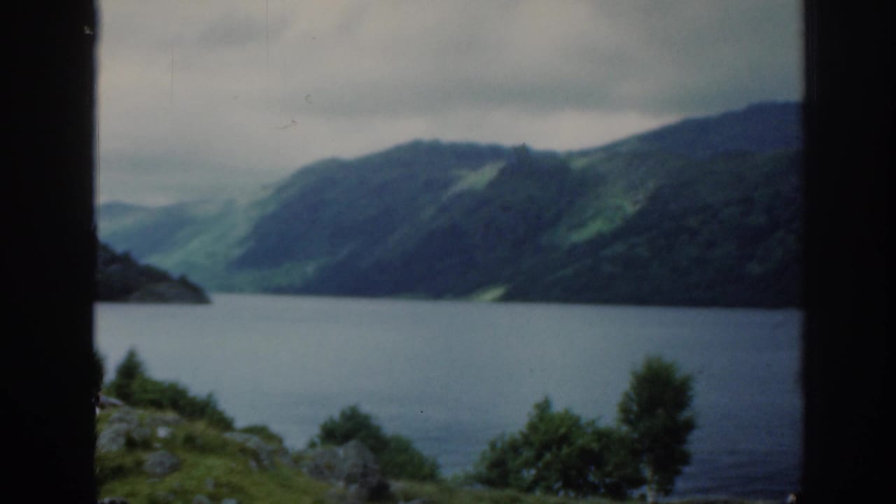 1961 Scotland home-movie still of a loch-like lake and green slopes, viewed through a dark frame with shrubs below.