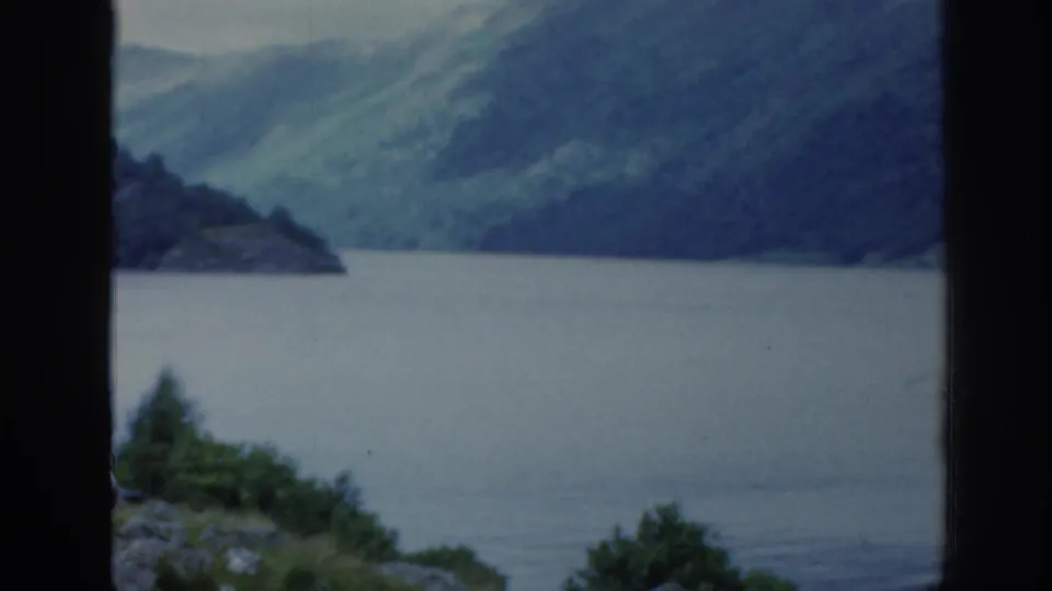 1961 Scotland 8mm still with muted blue-green lake water, steep hills, and a dark window edge crossing the view.