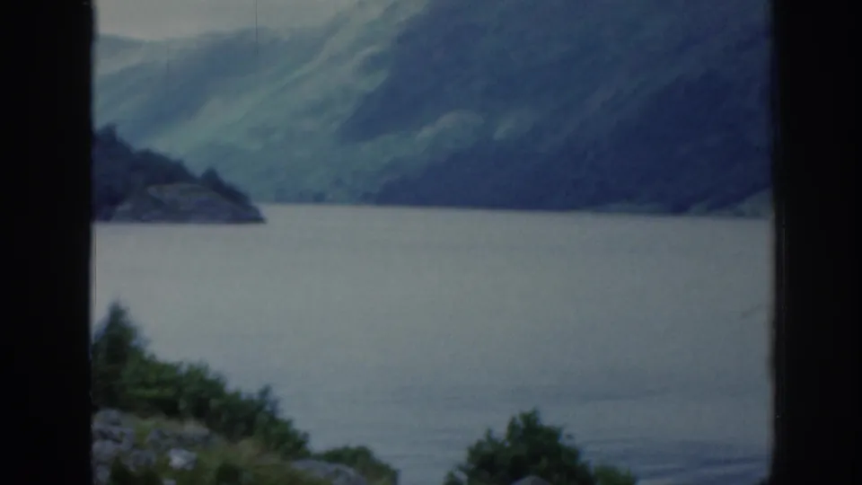 1961 Scotland home-movie frame showing calm water, distant green slopes, dark vertical framing, and low foreground vegetation.