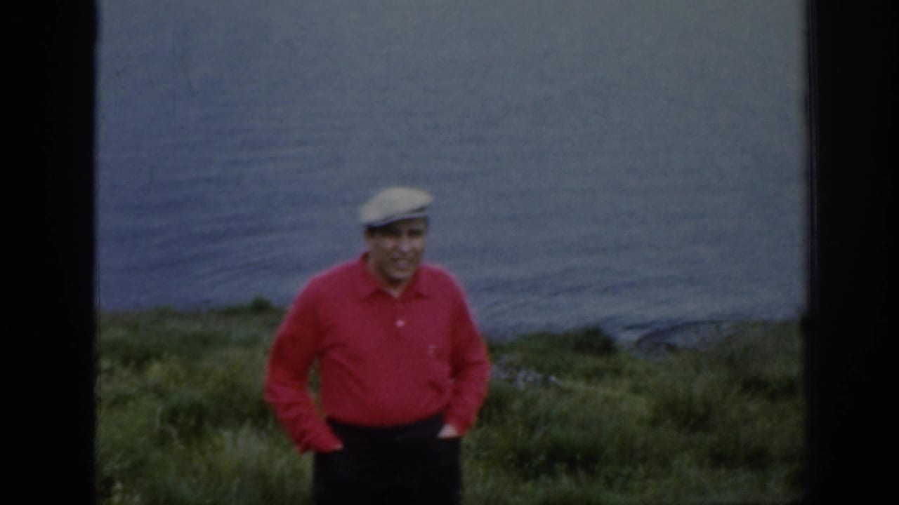 Man in a red shirt and light cap stands on a grassy hillside above a lake.