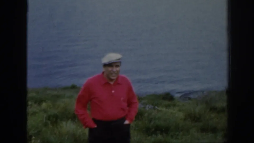 In Scotland, an adult man in a red shirt and light cap stands smiling on a grassy hillside with lake water behind him.