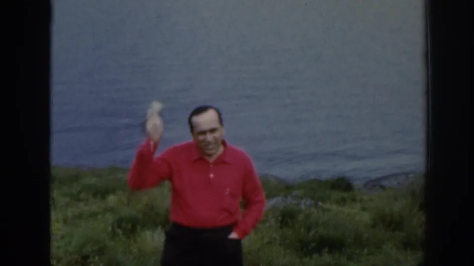 The adult man in a red shirt raises one hand in a wave while standing on a grassy slope above the Scottish lake.
