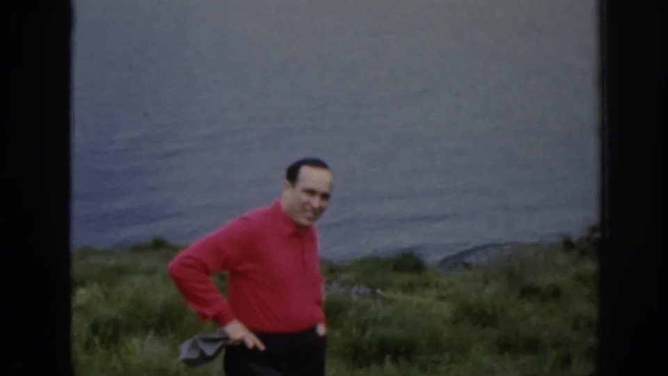 An adult man in a red shirt stands on the hillside with one hand on his hip, with still lake water and grass behind him.