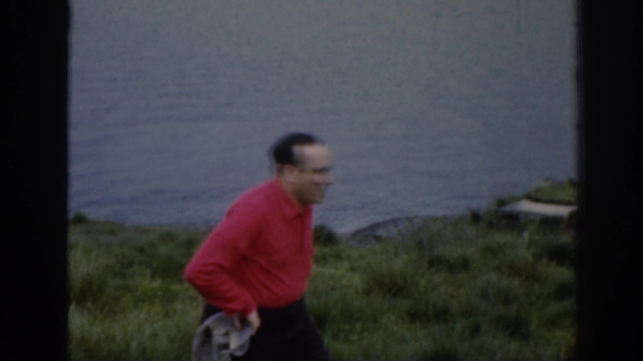 Man in a red shirt walking beside grassy vegetation with calm water behind him.