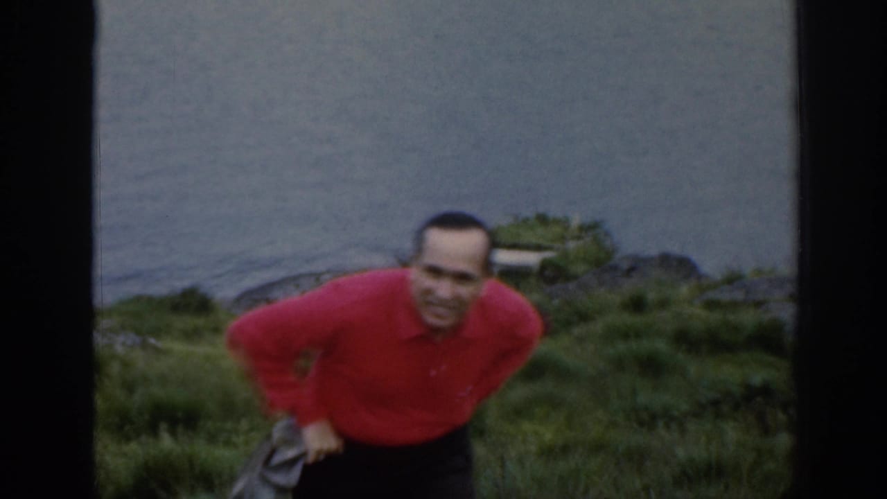 A smiling adult man in a red shirt moves toward the camera on a grassy Scottish waterside slope in 1961.