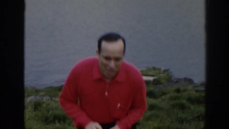 The man faces the camera from the grassy slope, with calm water and low vegetation behind him in this 1961 Scotland home-movie frame.