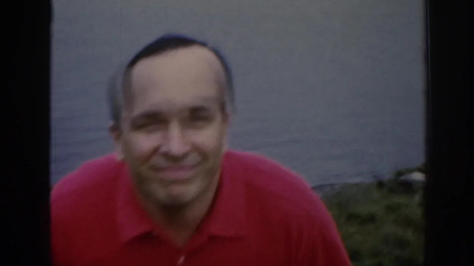 Close 8mm frame of a smiling adult man in a red shirt, filmed against water and grass in Scotland in 1961.