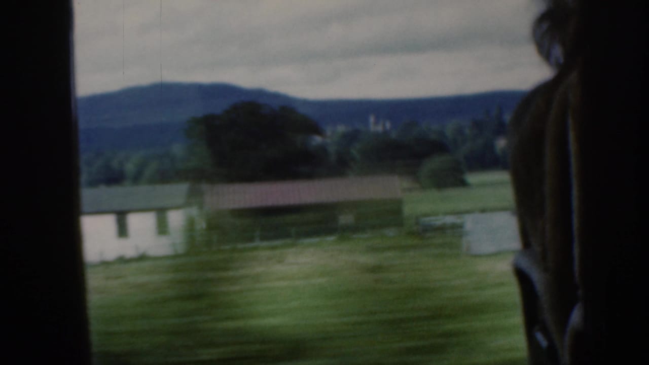 1961 Scotland 8mm train-window view showing green fields, a white farm building, distant hills, cloudy sky, and a passenger silhouette.