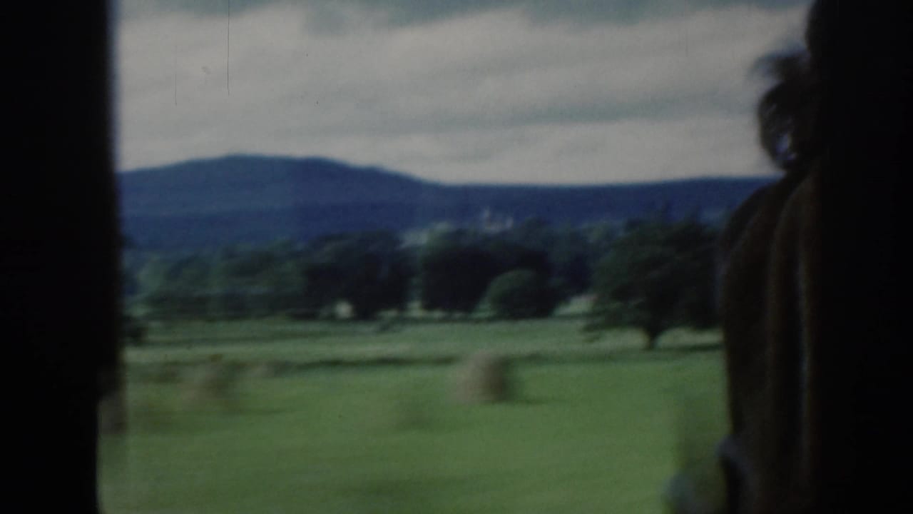 1961 Scotland countryside passes the train window, with green fields, tree lines, distant hills, cloudy sky, and a silhouetted passenger profile.