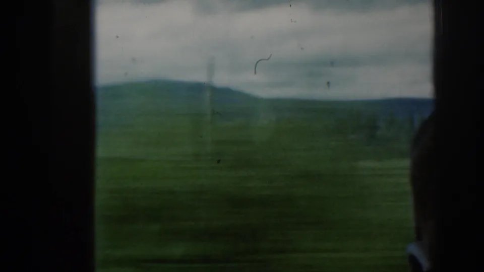 Soft 8mm frame from a moving train in Scotland, showing open fields, distant hills, cloudy sky, window framing, and a passenger silhouette.