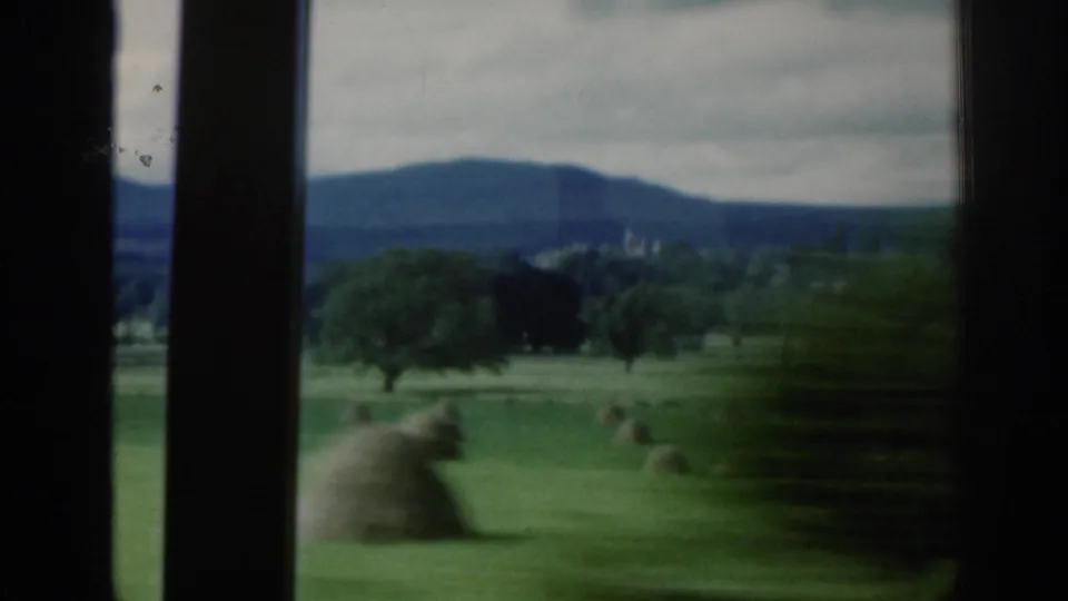 1961 Scotland home-movie view from a moving window across green fields, hay bales, trees, and distant hills under cloud.