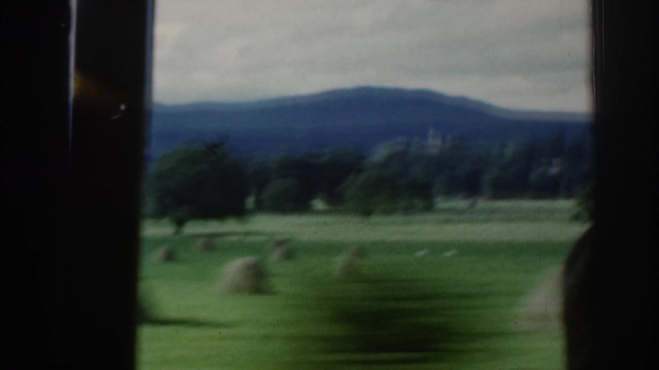 Window-framed countryside with green pasture, hay bales, trees, and hills beyond.