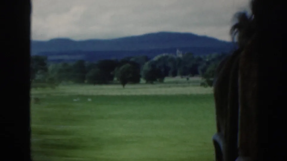Home-movie frame showing rural Scotland beyond a window, with a partially visible person close to the camera.
