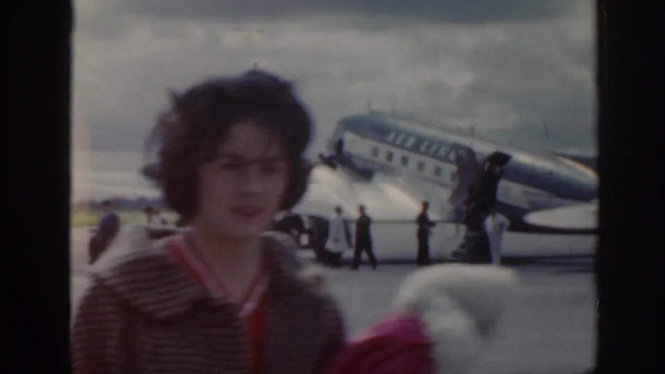 1961 Scotland airport tarmac scene with a young woman near the camera, people gathered, and a propeller airliner behind her.