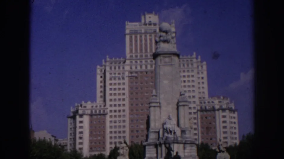 1966 Madrid home-movie frame showing a broad view of the monument and Edificio España with trees and sky behind it.