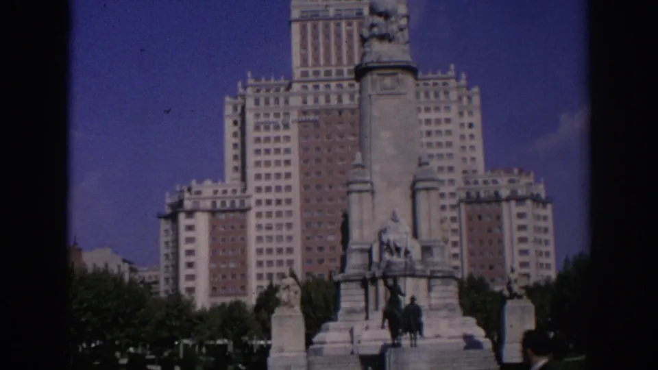 Madrid landmark view with the stone monument centered before Edificio España, framed by trees and dark 8mm gate edges.