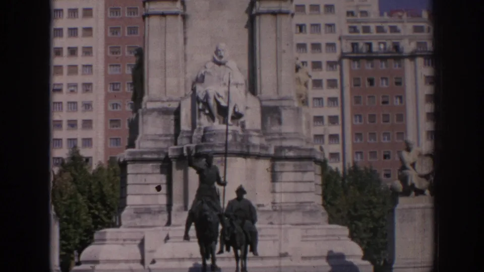 Home-movie frame from Madrid showing the monument base, two equestrian figures, trees, and multi-story buildings behind.