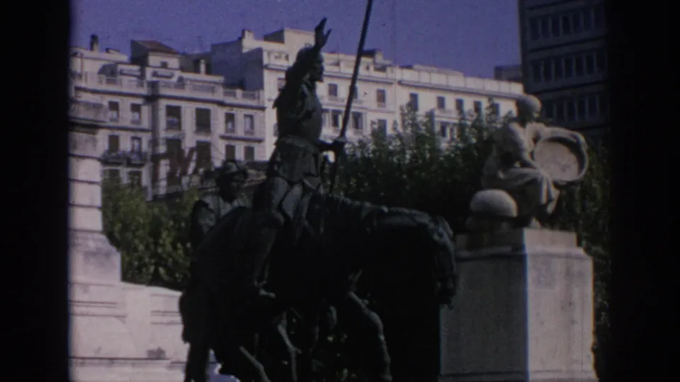 Visitor-shot 8mm view in Spain of an equestrian statue near a stone plinth, with trees and urban buildings in the background.