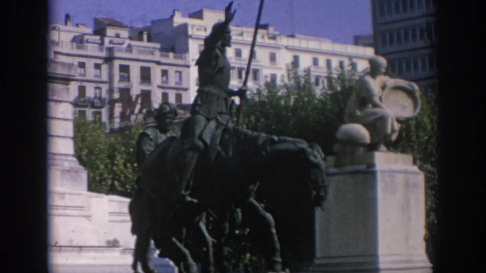 1960s 8mm frame from Spain showing a dark equestrian statue in front of pale monument stone and apartment-style facades.