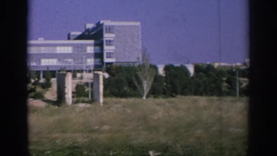 1966 Spain 8mm frame showing grass, trees, a fountain-like spray, and a modern building beyond an open urban area.