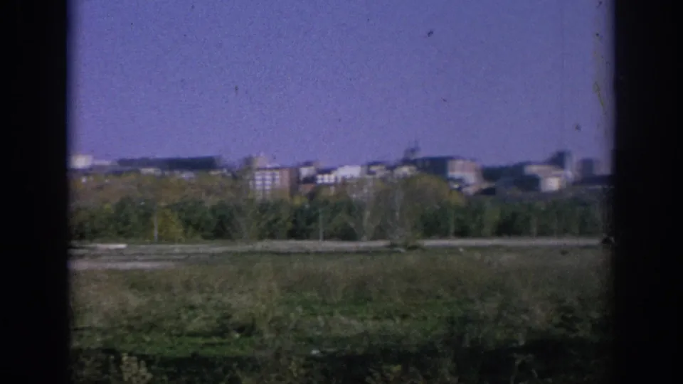 Spain 1966 8mm view of distant urban buildings beyond grass and roadside trees, with soft motion blur from travel filming.