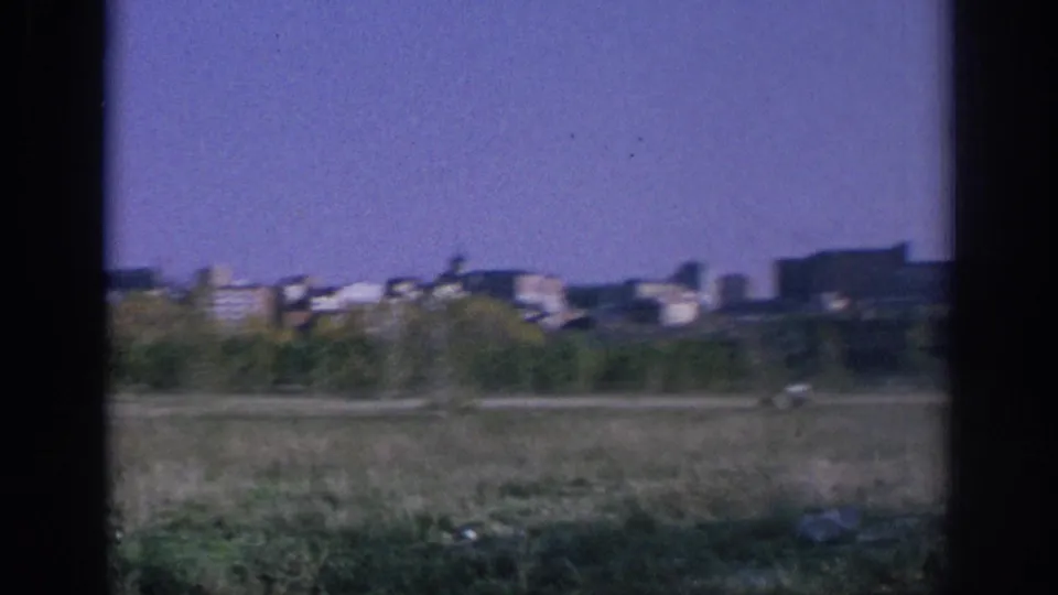 Spain 1966 frame showing a distant city skyline, low buildings, a small spire, and open grass in the foreground.