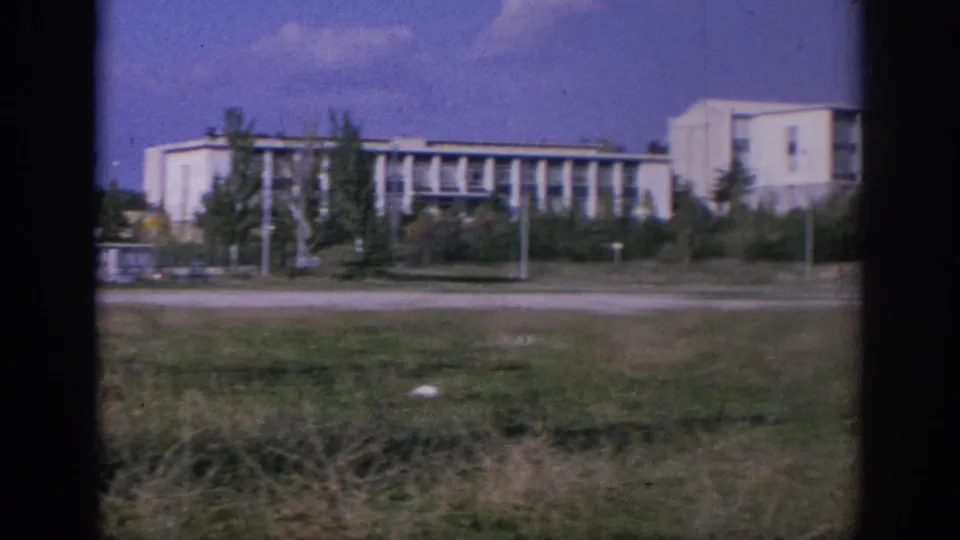 1966 Spain 8mm view of a long white institutional-looking building with repeating windows, trees, roadside grass, and dark window-frame edges.