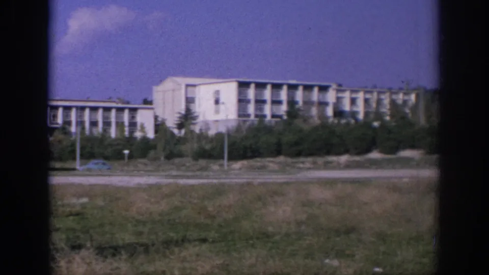 1966 Spain 8mm frame of a white flat-roofed building across an open field, with trees and roadway elements in front.