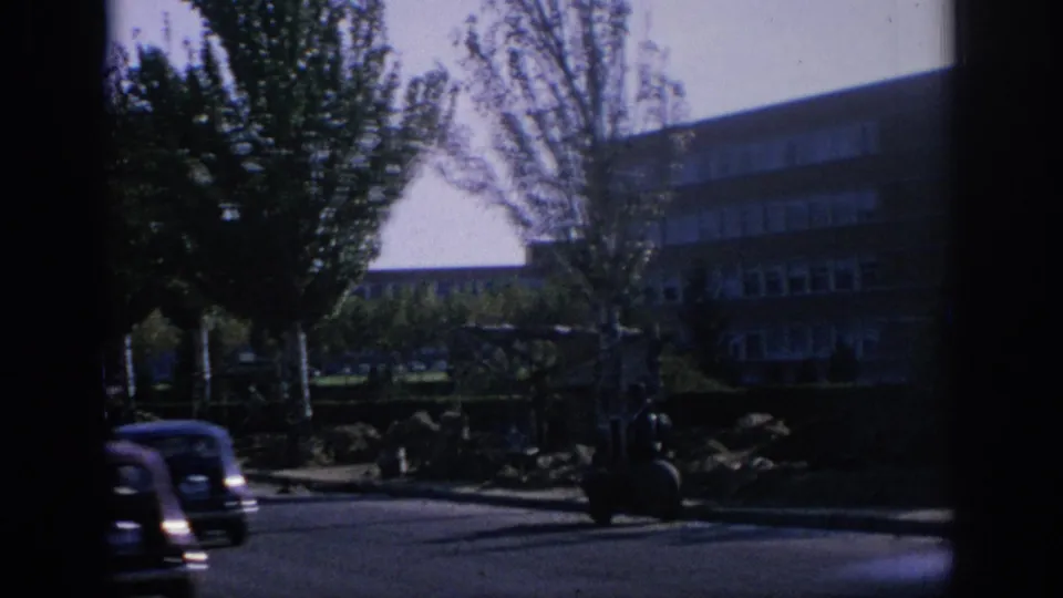1966 Spain 8mm frame showing a darker street-side view with trees, parked cars, and a multi-window building in shadow.