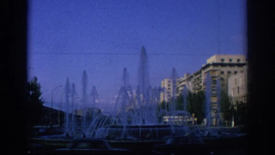 1966 Spain 8mm view of a public fountain with multiple water jets and apartment blocks beyond the plaza.