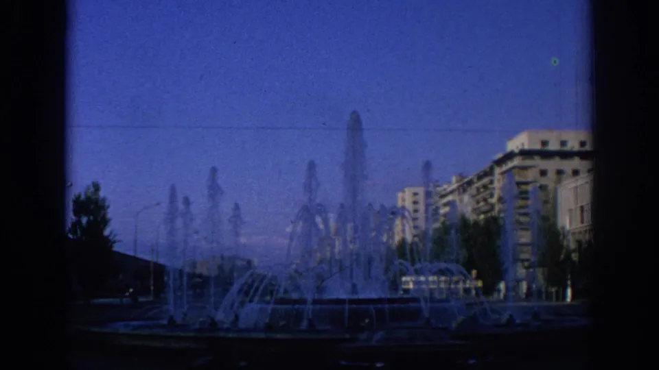 1966 Spain frame showing a working fountain, silhouetted foreground edges, and urban buildings under a blue sky.
