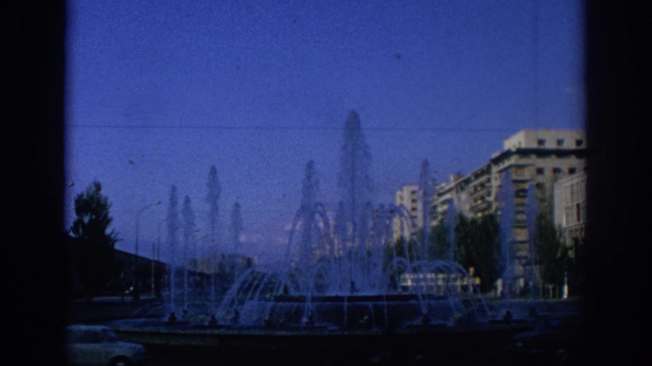 Large public fountain with water jets in front of apartment buildings and a blue sky.