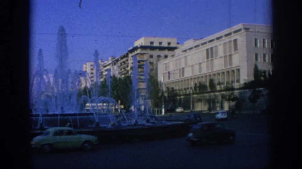 1966 Spain urban scene with a fountain in the foreground, classic cars on the street, and mid-century buildings behind.