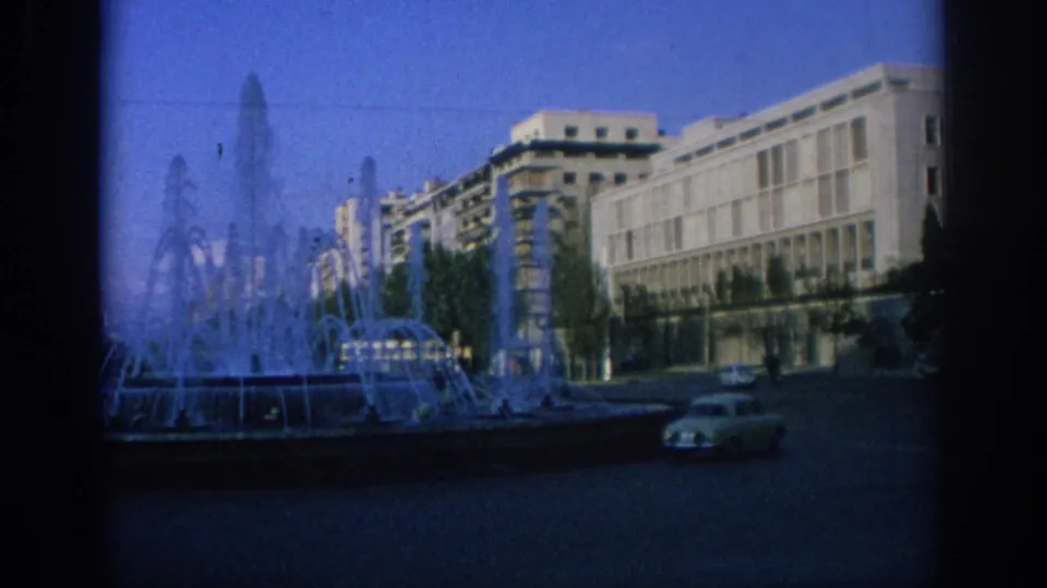 1966 Spain frame showing a public fountain, long modernist facade, trees, and 1960s cars crossing the foreground street.