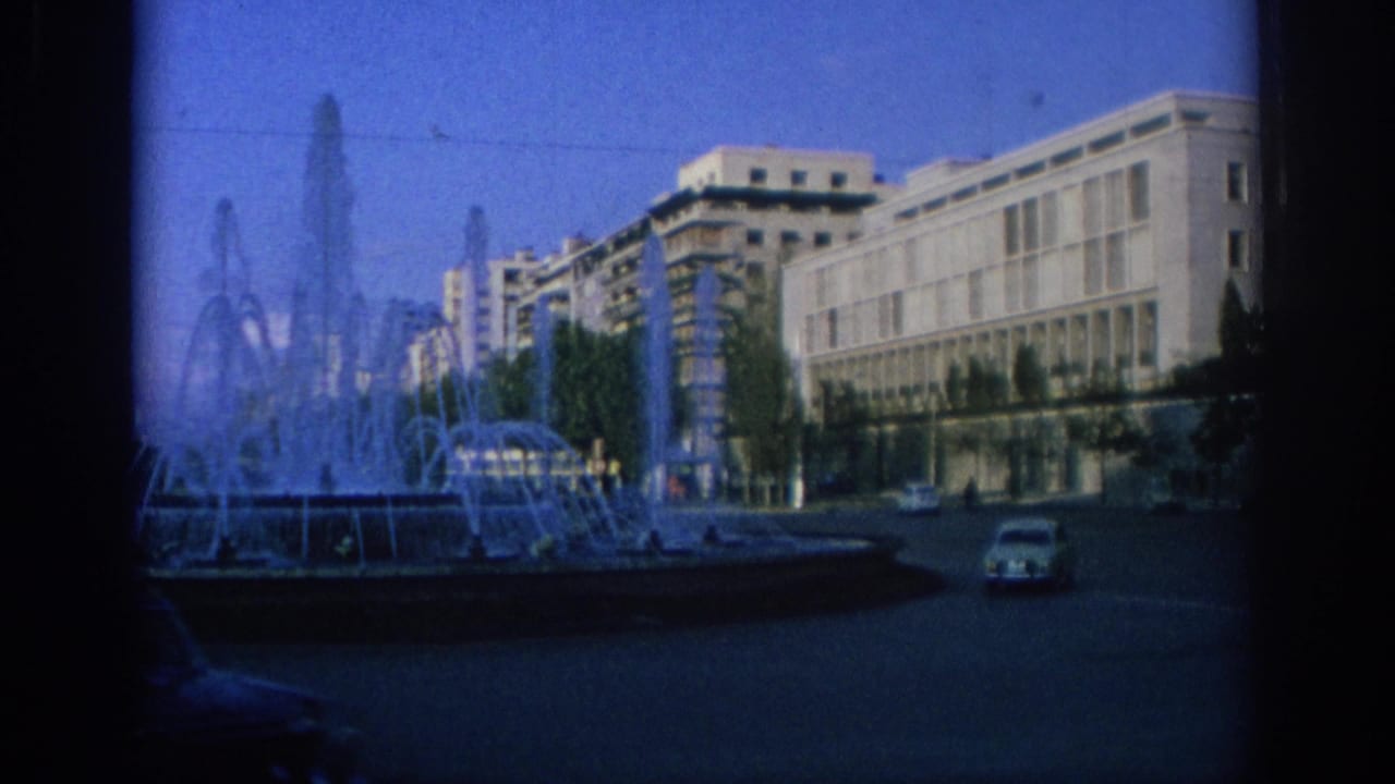 City square with fountain jets, pale modern building, historic facade, and a small car.