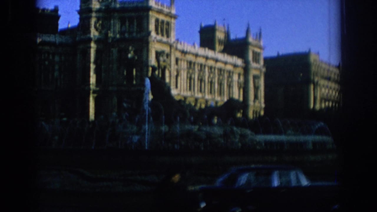 A grand urban facade appears behind a shadowed fountain in 1966 Spain, with a dark car partly visible along the street.