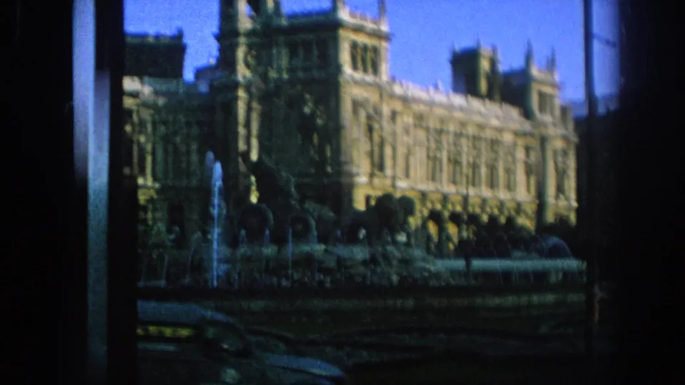 The handheld 8mm view holds on a large historic building and fountain sculpture in a Spanish city square.