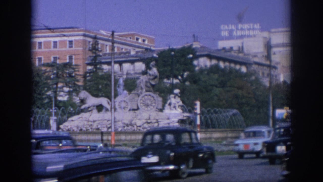 1966 Spain street view with classic cars moving past a large stone fountain and a CAJA POSTAL DE AHORROS sign in the background.