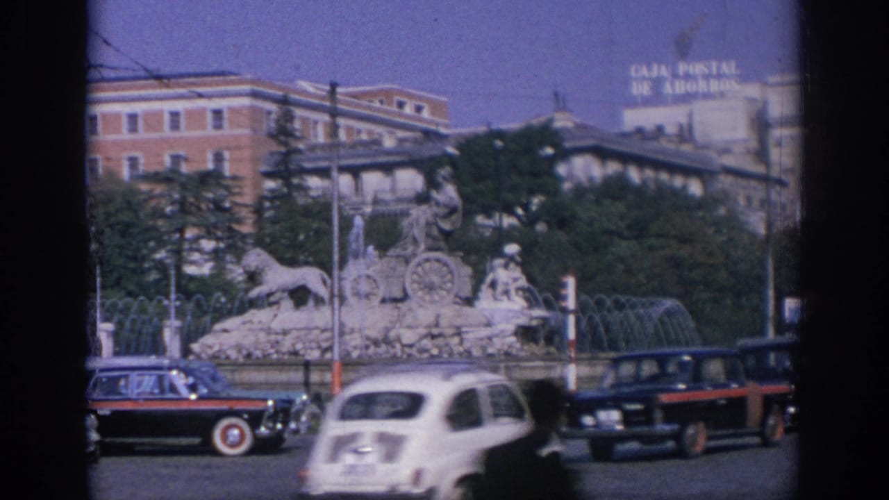 A white car and dark sedans cross in front of the fountain and traffic signal.