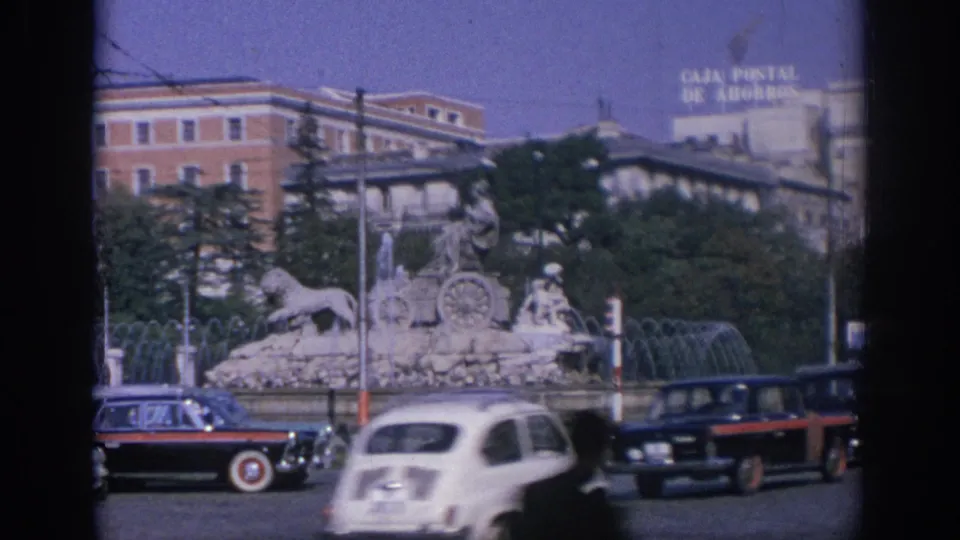 1966 Spain intersection with a white compact car, dark sedans, traffic signals, and the sculptural fountain behind them.