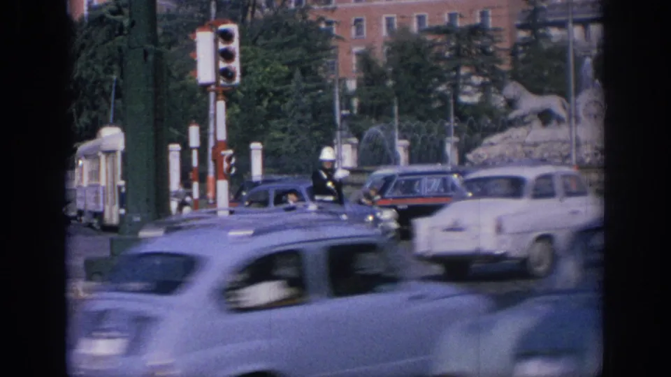 1966 Spain street scene with a white-helmeted traffic officer, passing cars, signals, and the fountain partly visible behind traffic.