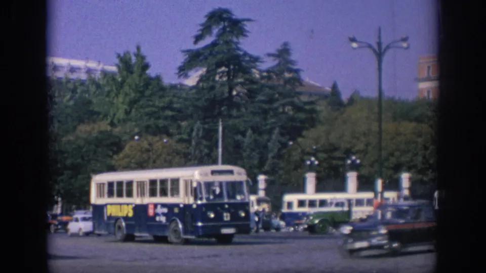 1966 Spain street scene with a Philips-branded bus, classic cars, a green vehicle, trees, and plaza lighting.