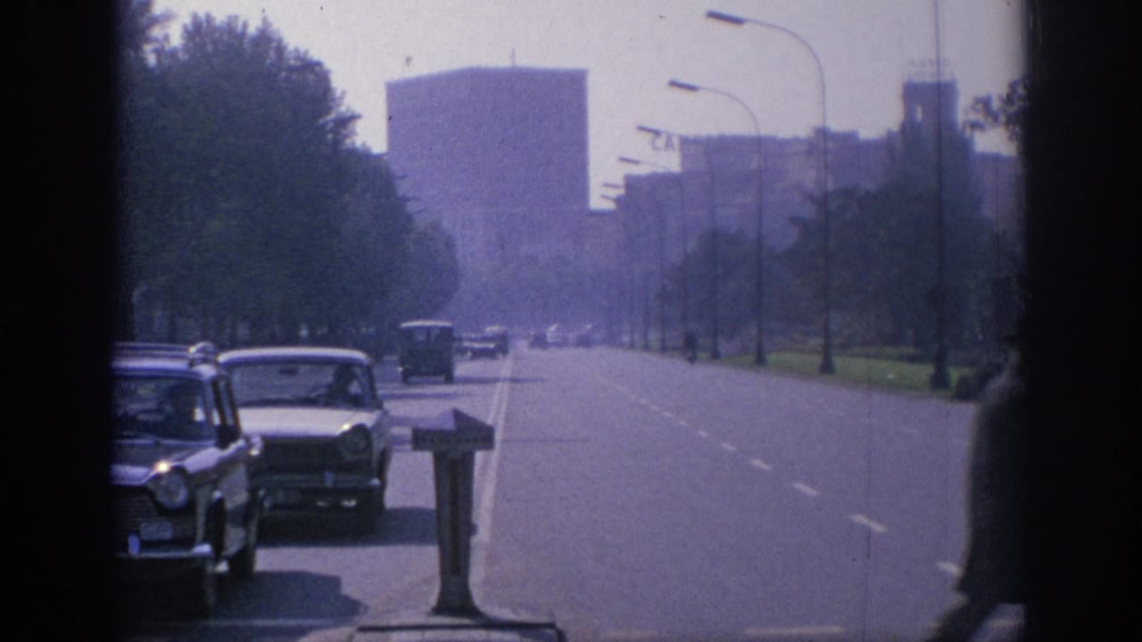 1966 Spain boulevard scene with classic cars, a distant bus or van, trees, streetlights, and large urban buildings in hazy 8mm color.