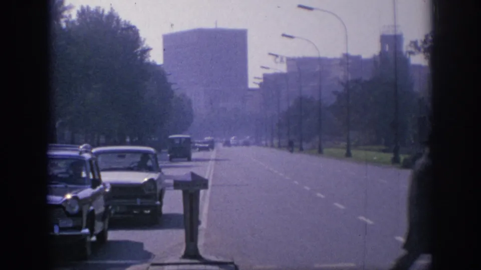 1966 Spain boulevard scene with classic cars, a distant bus or van, trees, streetlights, and large urban buildings in hazy 8mm color.