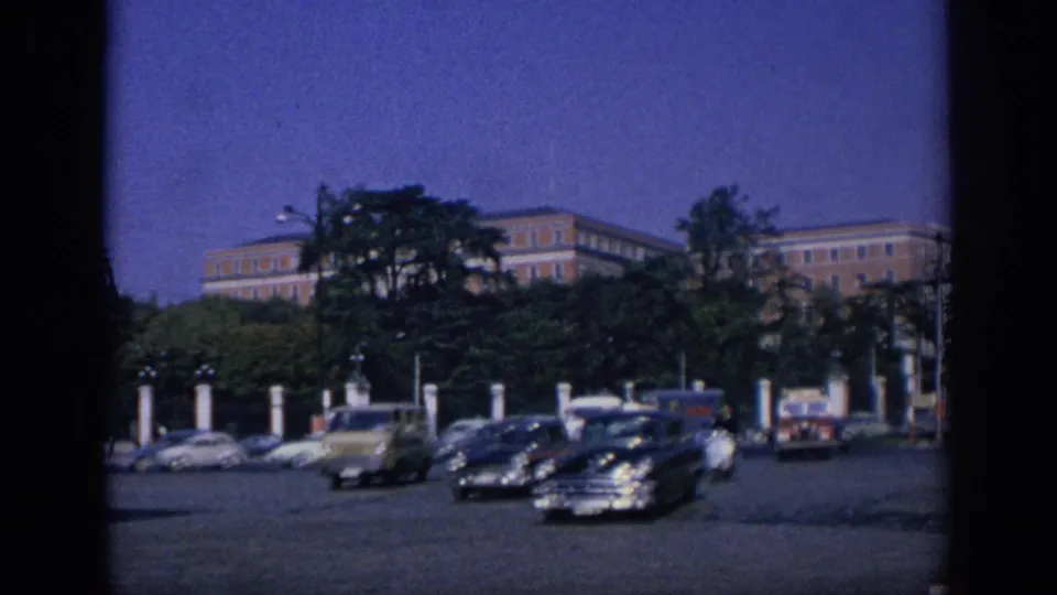 1966 Spain street traffic with several classic cars crossing the frame, trees, white posts, and a large building behind the greenery.