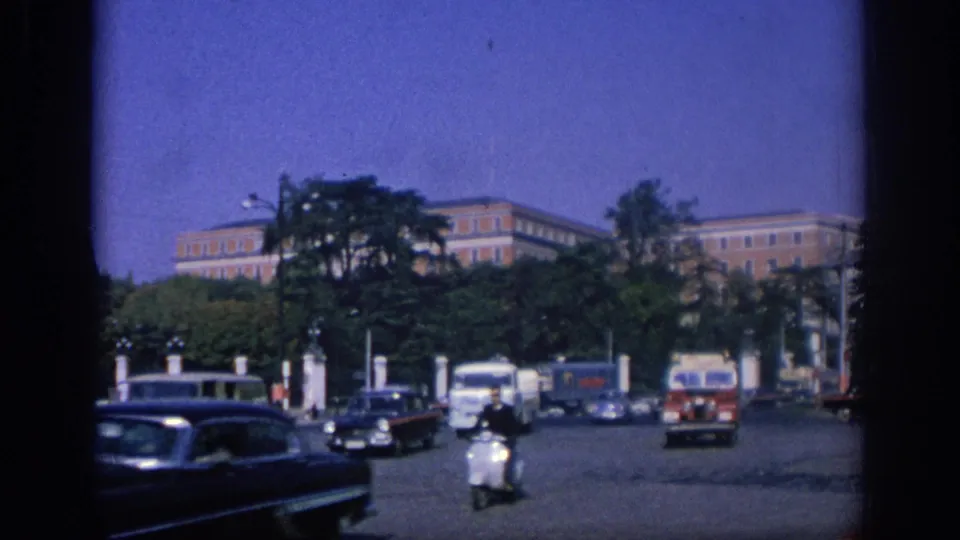 1966 Spain traffic scene with a scooter rider, classic cars, vans, trees, white posts, and large buildings under a blue sky.