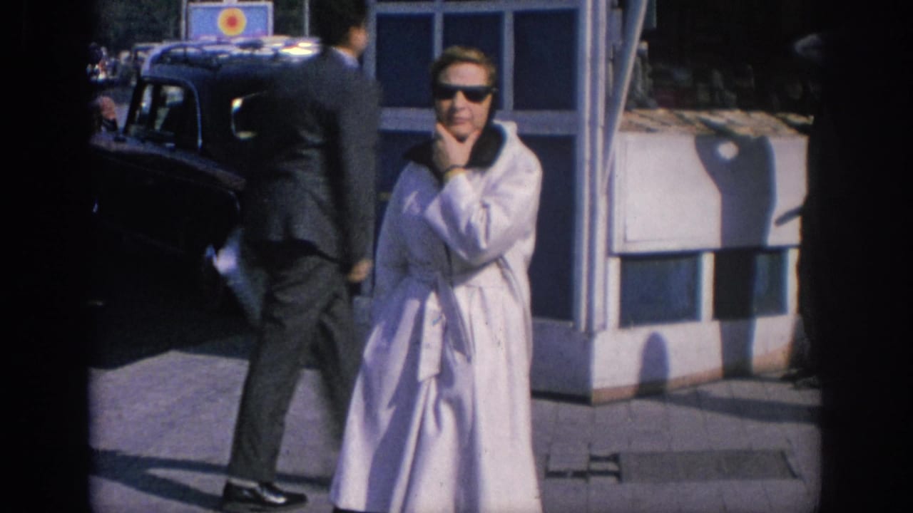 In Spain in 1966, a woman in a light raincoat and sunglasses stands near a black car while a pedestrian passes on the sidewalk.