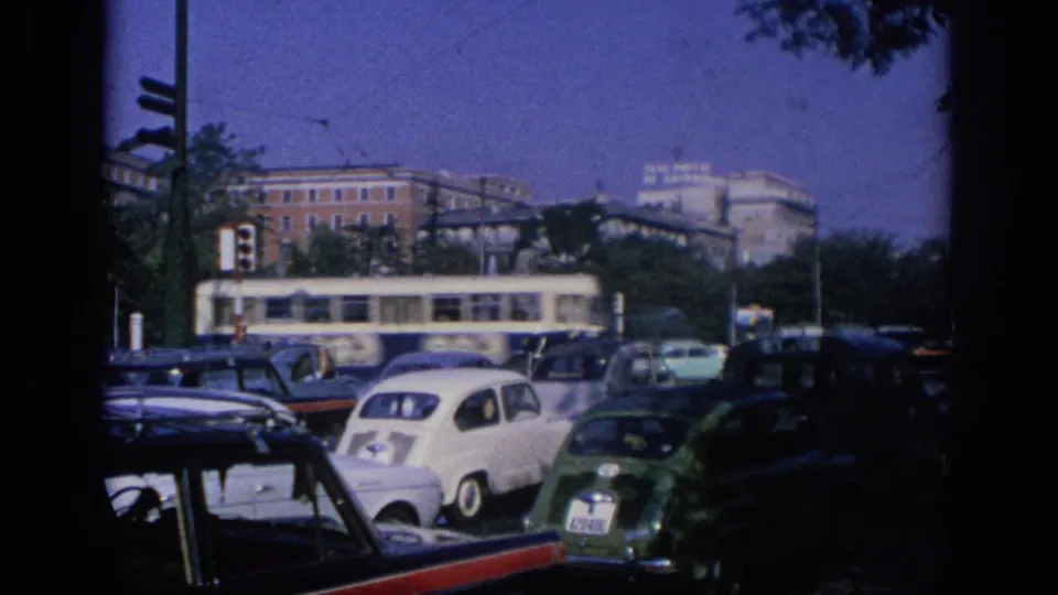 A 1966 Spain street view shows compact cars in traffic, a long transit vehicle, traffic lights, and large buildings in the background.