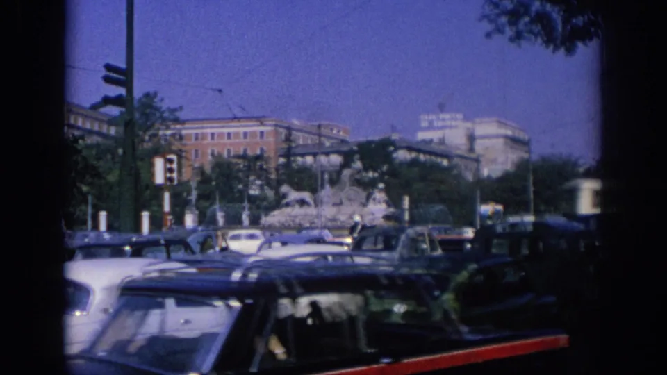 A Spain 1966 traffic scene shows cars clustered near a fountain, traffic signals, trees, and large urban buildings under a blue-purple sky.
